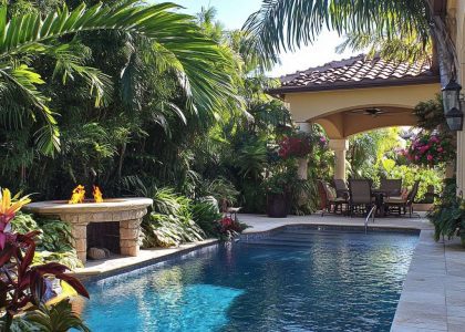 Backyard swimming pool glimmering under the sun, surrounded by creamy stone decking, lush greenery, vibrant flowers, and a cozy fire pit, creating a serene tropical oasis.